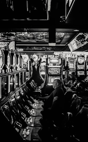A dimly lit casino filled with rows of slot machines, some occupied by people. The machines' lights contrast with the dark surroundings, creating a vibrant yet subdued atmosphere.