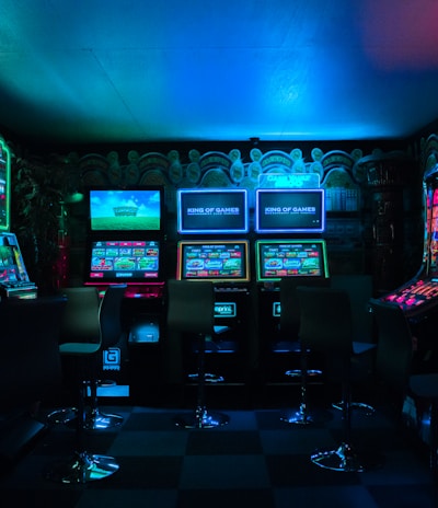 Dimly lit gaming arcade featuring a variety of brightly illuminated slot machines and video gaming screens. Neon signs and colorful displays create a vibrant atmosphere. The arrangement of gaming machines suggests an inviting space for entertainment, with chairs positioned for players.