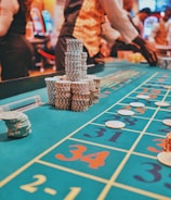 A casino scene with a roulette table features prominently, covered in green felt with colorful numbers and betting squares. Several stacks of poker chips are placed on different numbers, and the hands of several people are visible as they interact with the table. In the background, there are slot machines and more people, creating a vibrant and busy atmosphere.