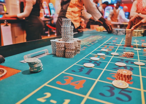 A casino scene with a roulette table features prominently, covered in green felt with colorful numbers and betting squares. Several stacks of poker chips are placed on different numbers, and the hands of several people are visible as they interact with the table. In the background, there are slot machines and more people, creating a vibrant and busy atmosphere.