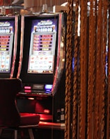 A pair of casino slot machines, each with detailed screens displaying various game outcomes and jackpot amounts. The chairs in front of the slot machines are dark and cushioned for comfort. The background features a hanging curtain made of golden ropes and other casino decorations are visible.