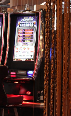 A pair of casino slot machines, each with detailed screens displaying various game outcomes and jackpot amounts. The chairs in front of the slot machines are dark and cushioned for comfort. The background features a hanging curtain made of golden ropes and other casino decorations are visible.