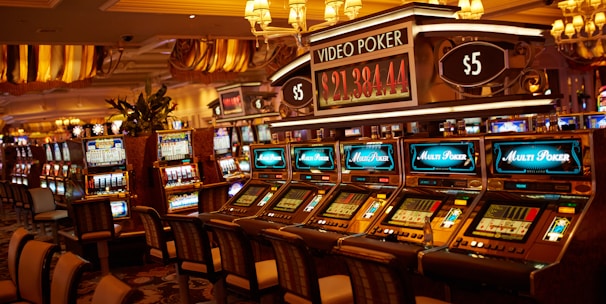 A casino floor featuring rows of brightly lit video poker machines. The area is illuminated by elegant chandeliers, with a display board showing a large sum of money, suggesting a possible jackpot. The rich decor includes luxurious carpets and comfortable seating, adding to the opulent atmosphere.