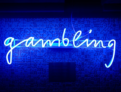 A bright blue neon sign spells out the word 'gambling' in cursive script. The background features a textured wall covered in domino tiles, also illuminated by the blue light, creating a vivid and visually striking scene.