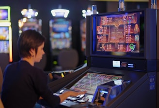 A person is seated at a slot machine console in a casino, focusing intently on the game displayed on the screen. The background shows several other illuminated gaming machines, adding a vibrant and colorful atmosphere to the scene.