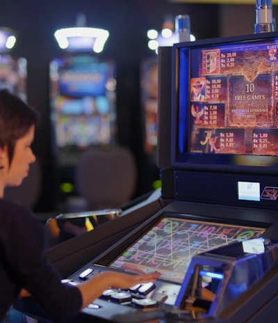 A person is seated at a slot machine console in a casino, focusing intently on the game displayed on the screen. The background shows several other illuminated gaming machines, adding a vibrant and colorful atmosphere to the scene.