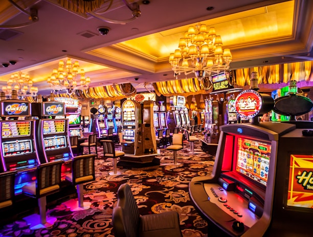 A vibrant casino interior filled with rows of colorful slot machines, each illuminated with bright lights and displays. The ceiling is adorned with elegant chandeliers and golden drapes, creating a luxurious atmosphere. The patterned carpet adds to the elaborate design of the space.