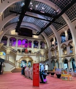 The interior of a large, ornately designed building with arched ceilings and intricate architectural details. The space features gaming machines lined up along the floor, under a high ceiling with a grid pattern. Lighting in purple and white tones enhances the ambiance.