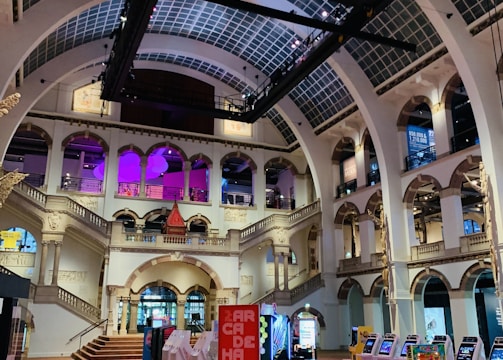 The interior of a large, ornately designed building with arched ceilings and intricate architectural details. The space features gaming machines lined up along the floor, under a high ceiling with a grid pattern. Lighting in purple and white tones enhances the ambiance.