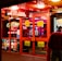 A brightly lit entrance to a casino with neon signs, including one advertising a £500 jackpot. The doors have clear signage indicating entry is restricted to those over 18. A person in a red jacket stands to the right, possibly interacting with a machine or display.