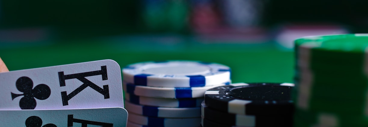 A casino scene featuring two playing cards, the ace and king of clubs, partially concealed by a player's hand. Nearby, there are stacks of multicolored poker chips on a green felt table, with blurred colorful background elements suggesting other chip stacks or cards.