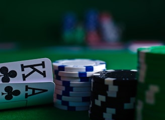 A casino scene featuring two playing cards, the ace and king of clubs, partially concealed by a player's hand. Nearby, there are stacks of multicolored poker chips on a green felt table, with blurred colorful background elements suggesting other chip stacks or cards.
