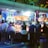 A group of people standing in front of an illuminated amusement arcade, engaging with various gaming machines. The environment is bustling with activity, and colorful lights create a vibrant atmosphere. The ceiling is decorated with large, playful images of dice and other game-related graphics, enhancing the lively mood.
