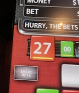 A digital display screen showing a casino betting interface, with text prompting to hurry bets and a highlighted number 27 in red. There are options for betting amounts and a 'share bets' button.