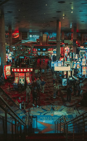 A bustling casino floor filled with groups of slot machines and people walking around. The atmosphere is vibrant with neon lights and illuminated signs. The interior has a ceiling supported by large pillars and there are stairs leading down to the gaming area.