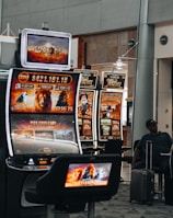 A collection of slot machines is situated inside a building with bright, colorful screens displaying various games and gaming themes. A person is seated nearby, seemingly relaxed with luggage beside them, and the atmosphere is lively with electronic displays.