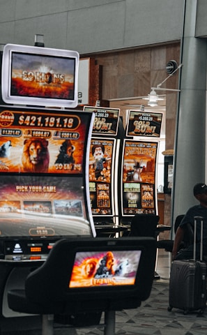 A collection of slot machines is situated inside a building with bright, colorful screens displaying various games and gaming themes. A person is seated nearby, seemingly relaxed with luggage beside them, and the atmosphere is lively with electronic displays.